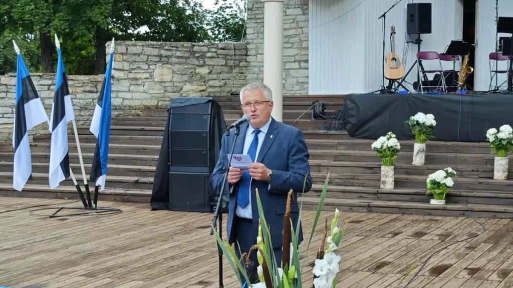Prillidega mees ülikonnas peab puidust laval mikrofoni käes hoides kõnet, taustal Eesti lipud.