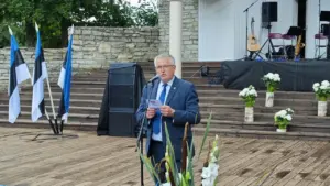 Prillidega mees ülikonnas peab puidust laval mikrofoni käes hoides kõnet, taustal Eesti lipud.